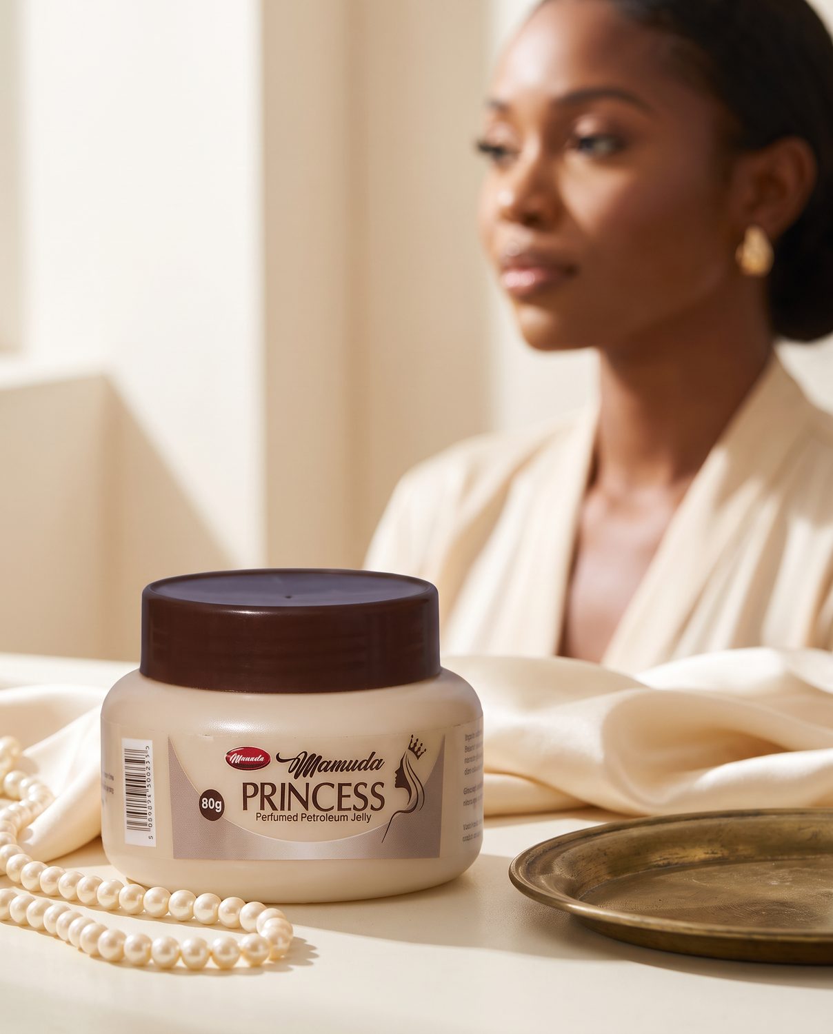 The beige and brown Mamuda Princess perfumed petroleum jelly jar large in the foreground on a cream surface, with an elegantly dressed Nigerian woman softly out of focus behind