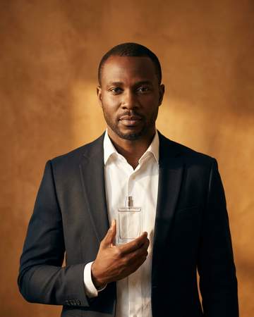 A Nigerian man in a white shirt and dark tailoring holding a clear glass fragrance bottle