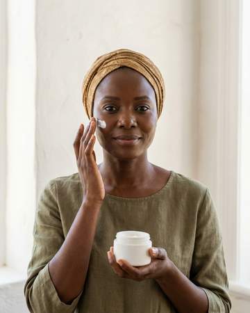 A Nigerian woman in an ochre headwrap applying cream to her cheekbone, holding a plain white jar